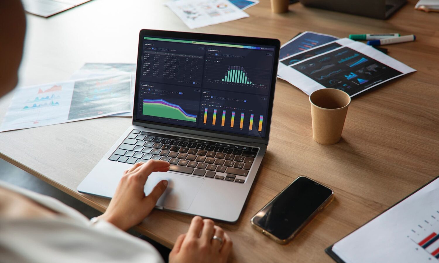 Data analyst working on laptop displaying colorful charts and graphs. Desk scattered with financial reports, smartphone, and coffee cup, showcasing modern business process analysis and performance tracking