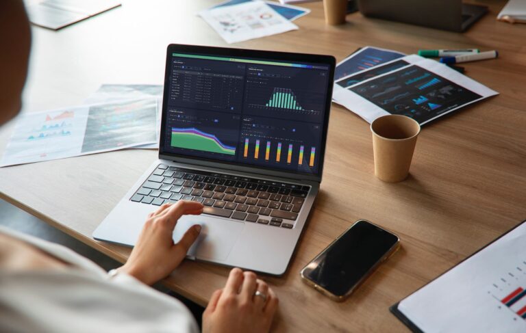 Data analyst working on laptop displaying colorful charts and graphs. Desk scattered with financial reports, smartphone, and coffee cup, showcasing modern business process analysis and performance tracking